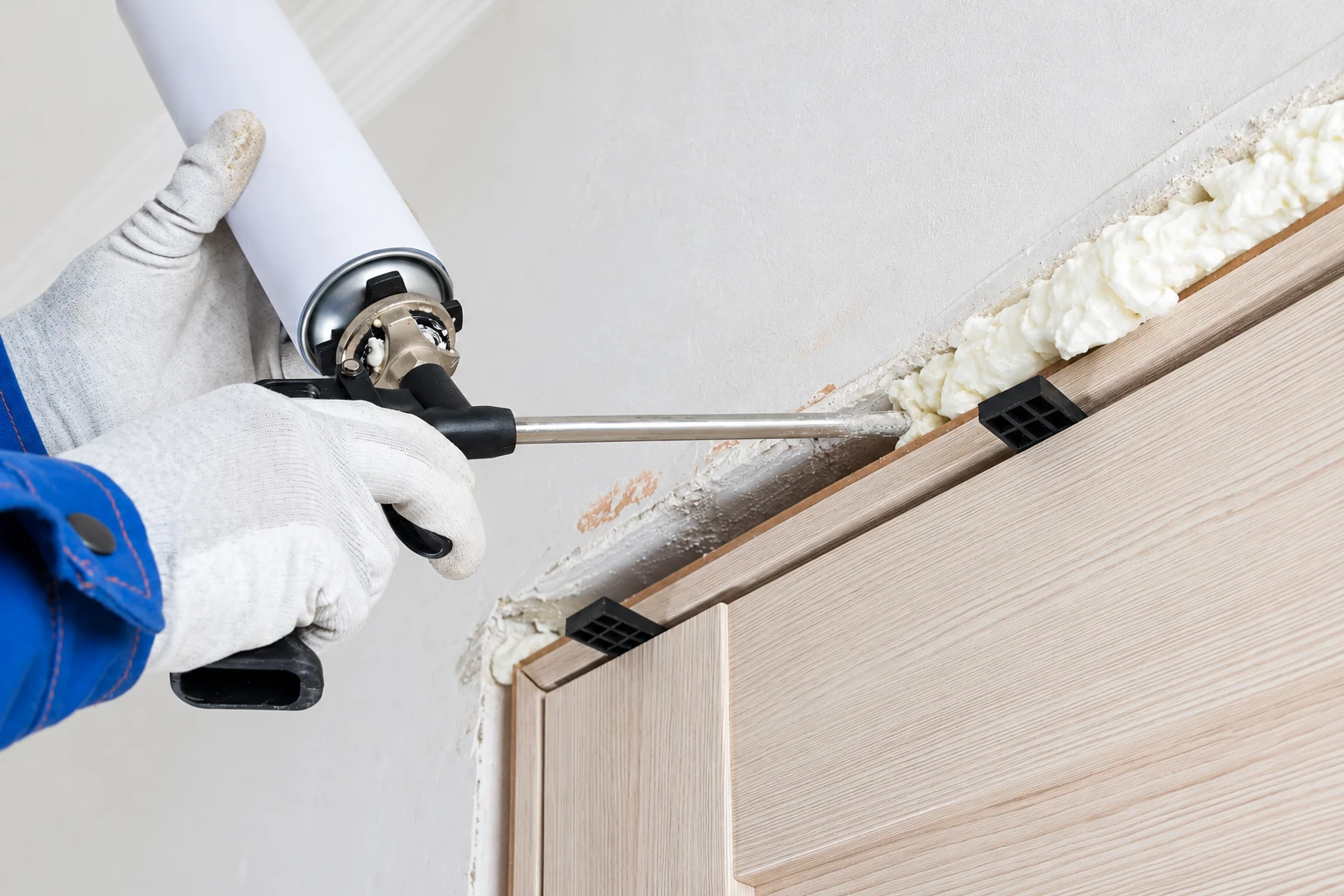 Gloved worker applies expanding foam sealant along the gap beside a wooden door frame.