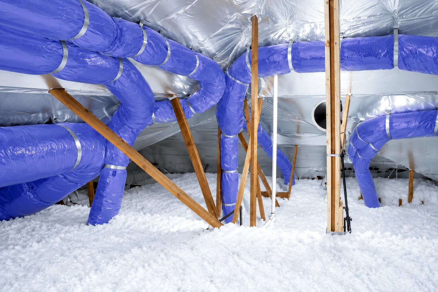 Attic filled with loose white blown-in insulation beneath reflective foil, with large blue ducts running between wooden supports.