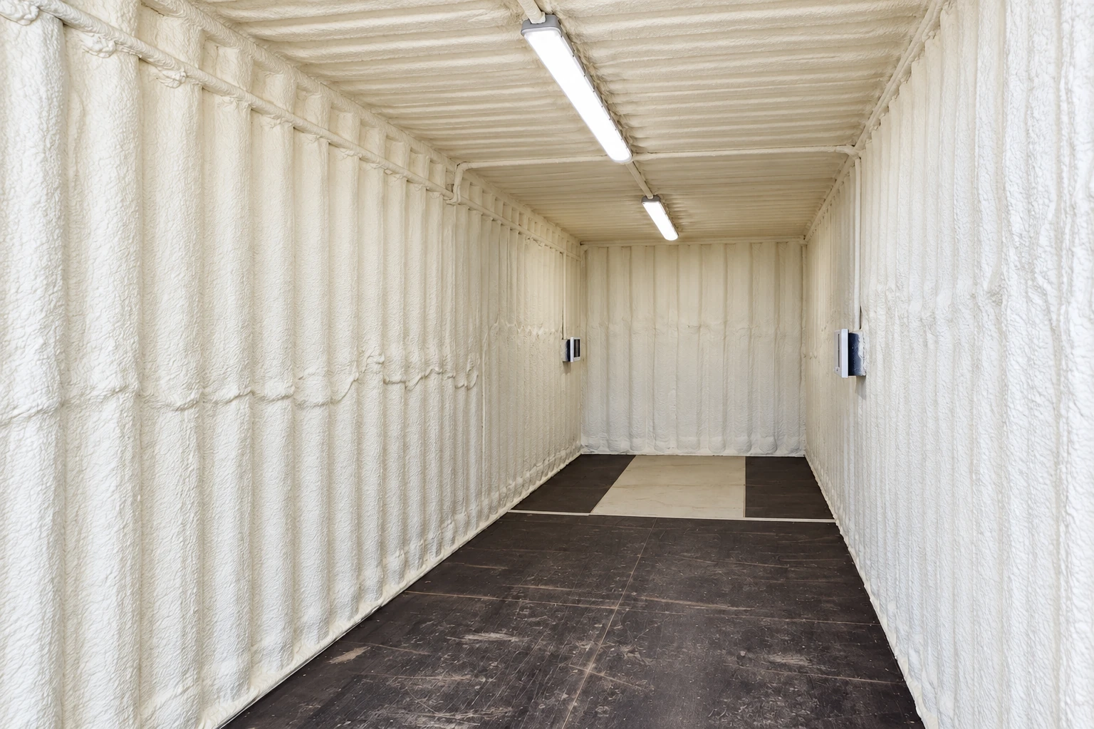 A narrow commercial interior is coated with white spray foam across its walls and ceiling, with a dark tiled floor and overhead lights.
