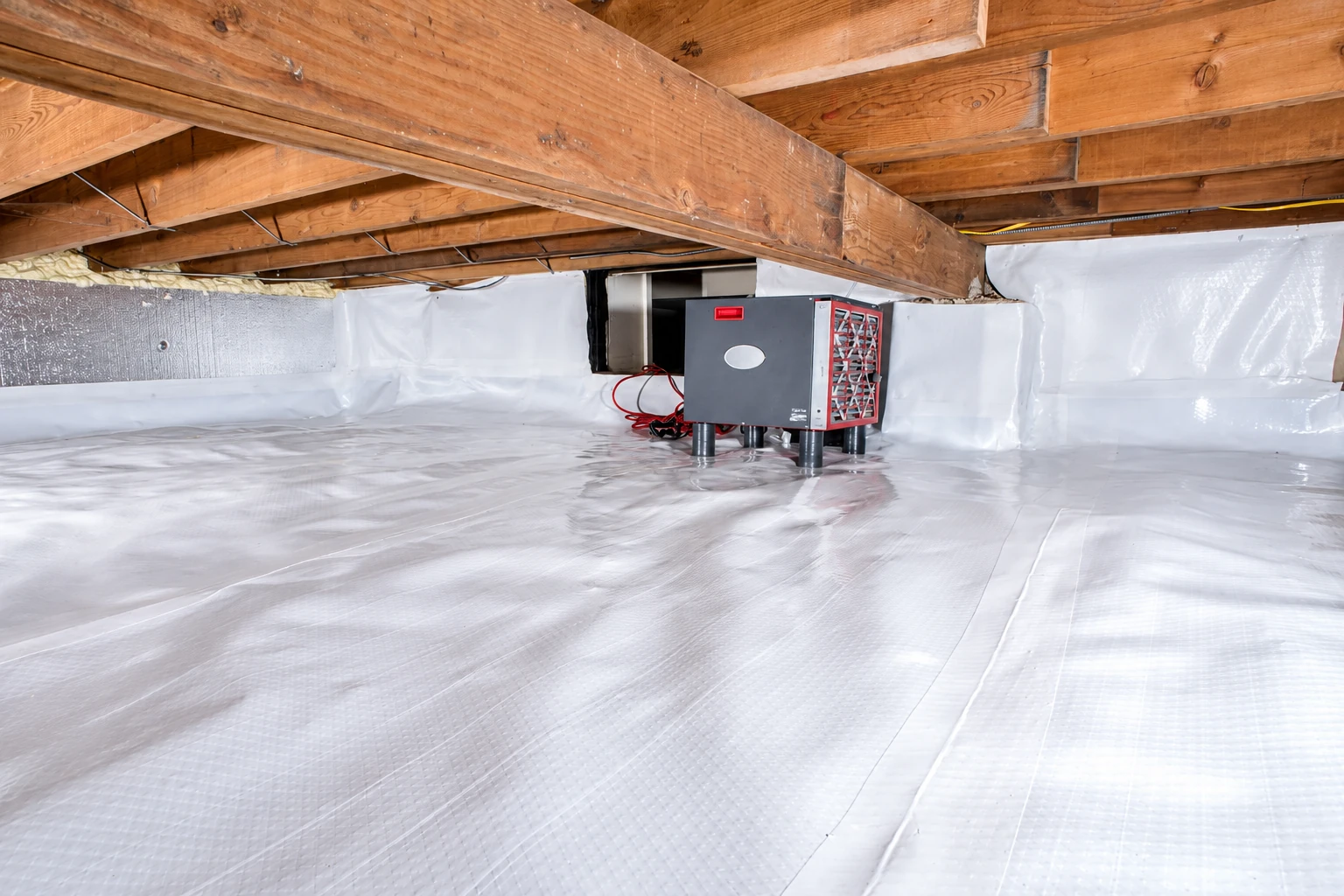 White vapor barrier covers a crawl-space floor and walls around a small dehumidifier beneath wooden joists.